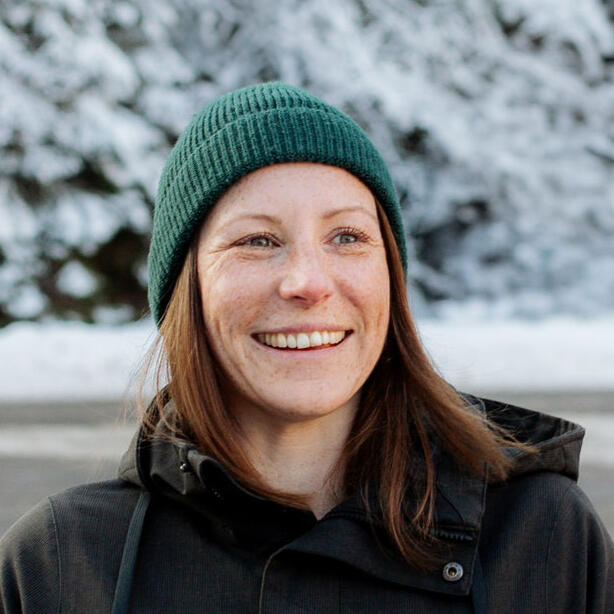 Close-up of Jen smiling in a green beanie and dark jacket, with snow-covered trees behind her.