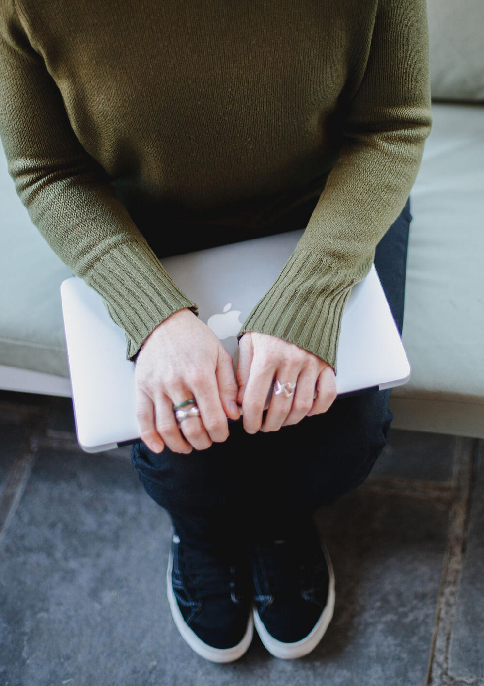 Top-down view of Jen in an olive-green knitted jumper and black trainers, hands resting on a closed laptop on her lap.