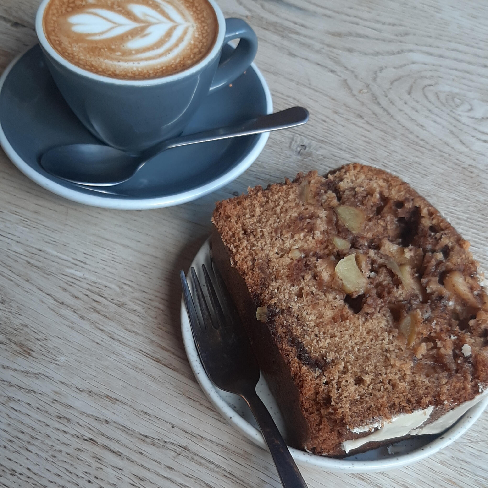 Meeting essentials: a flat white in a dark blue cup and saucer, next to a thick slice of apple cinnamon cake. Side-profile image of Jen next to splitboard tracks in the snow. She's smiling, wearing black clothing, a grey beanie, snowboard goggles with a pink strap, and a backpack. Snow-covered mountains and a partly cloudy sky are in the background.
