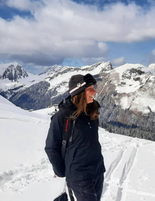 Side-profile image of Jen next to splitboard tracks in the snow. She's smiling, wearing black clothing, a grey beanie, snowboard goggles with a pink strap, and a backpack. Snow-covered mountains and a partly cloudy sky are in the background.
